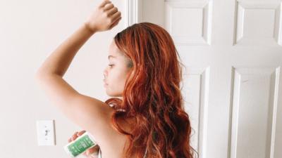 Woman with dyed red long hair with her left arm up applying Schmidt's Jasmine Tea Sensitive Skin natural deodorant facing a bathroom door